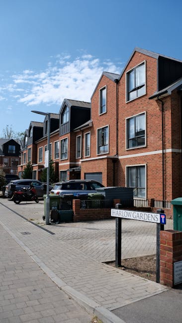 A modern residential street featuring multi-story brick townhouses with large windows and pitched roofs under a clear blue sky. In the foreground, a paved sidewalk runs alongside the houses, with neat