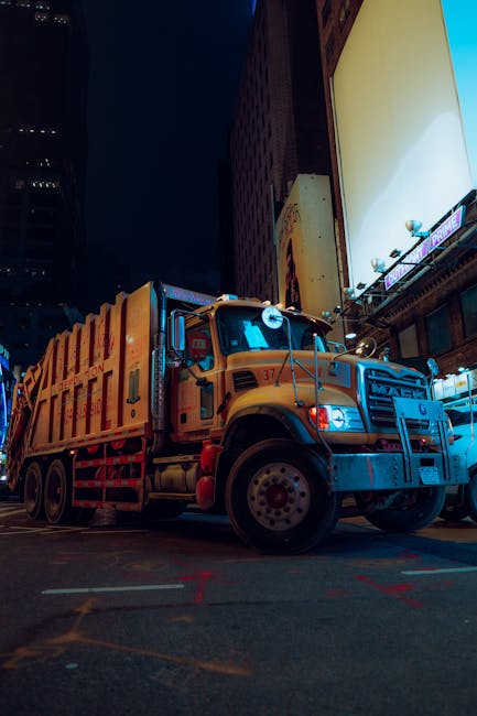 The image shows a large yellow street cleaning or waste collection truck parked on a city street at night, illuminated by nearby building lights and billboards. Tall buildings with illuminated windows
