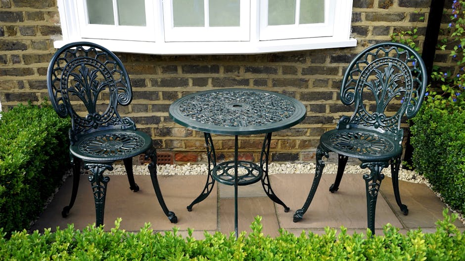 A small garden patio featuring two ornate black metal chairs with intricate floral and scrollwork designs, positioned on either side of a matching round metal table with a decorative patterned surface